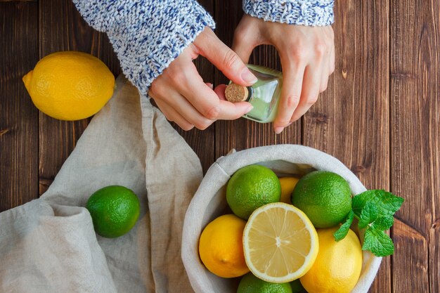 Photo gratuite vue de dessus citrons dans le panier avec un chiffon blanc, mains tenant une bouteille de jus de citron sur une surface en bois. verticale