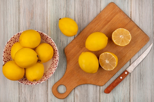 Vue de dessus de citrons entiers de forme arrondie sur un bol avec des citrons isolés sur une planche de cuisine en bois avec un couteau sur un mur en bois gris