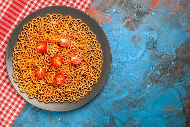 Vue de dessus des coeurs de pâtes italiennes coupés des tomates cerises sur une assiette ovale sur une nappe à carreaux rouges et blancs sur une table bleue avec un espace libre