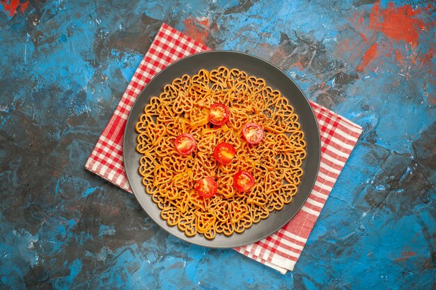 Vue de dessus des coeurs de pâtes italiennes coupés des tomates cerises sur une assiette sur un torchon à carreaux blanc rouge sur une table bleue