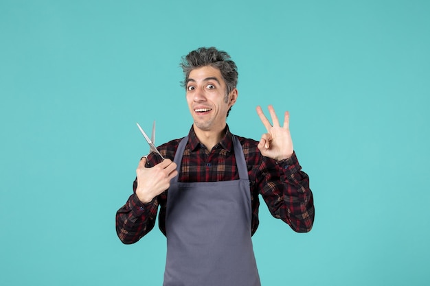 Vue de dessus d'un coiffeur masculin souriant portant un tablier gris et tenant des ciseaux montrant trois sur fond bleu de couleur douce