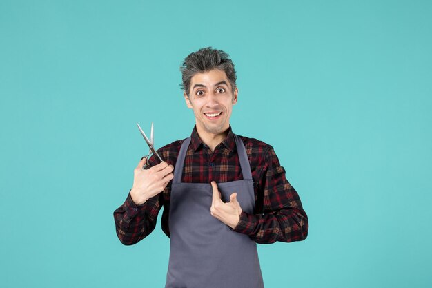 Vue de dessus d'un coiffeur masculin surpris portant un tablier gris et tenant des ciseaux se pointant sur un fond de couleur bleu doux