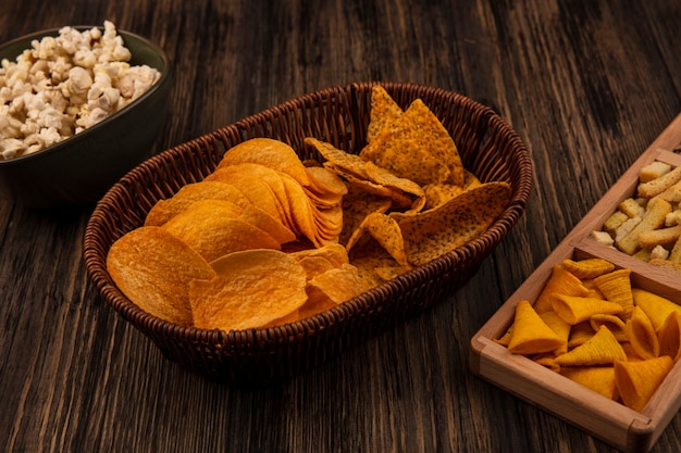 Vue de dessus des collations de maïs en forme de cône croustillant sur une plaque divisée en bois avec des graines de tournesol décortiquées avec des chips épicées sur un seau avec des pop-corn sur un bol sur une table en bois