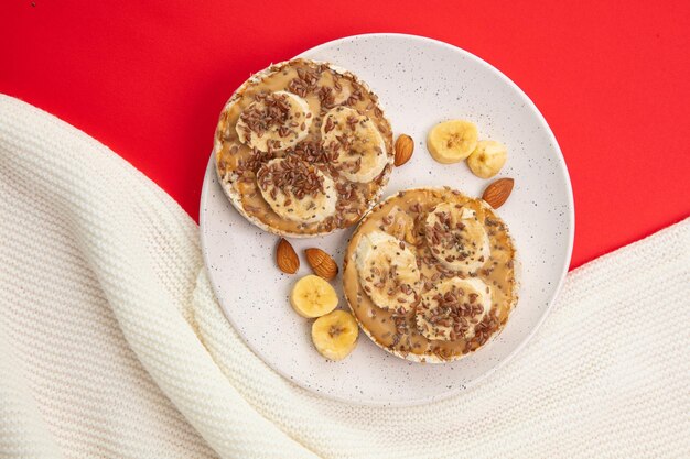 Vue de dessus des collations de pain croustillant au beurre d'arachide et à la banane avec des tranches d'amande et de banane dans une assiette sur un tissu blanc sur fond rouge