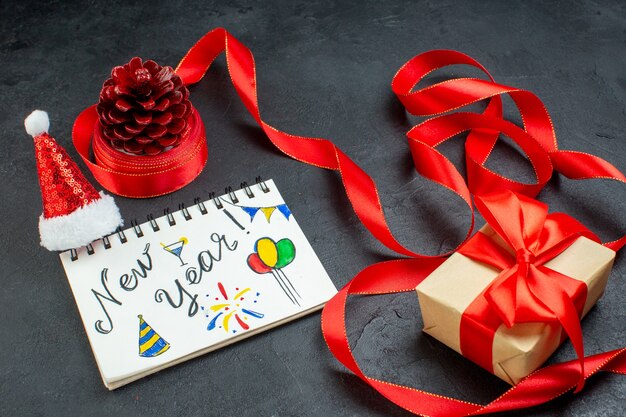 Vue de dessus d'un cône de conifère cadeau avec ruban rouge et cahier avec écriture de nouvel an et chapeau de père Noël beau cadeau sur fond sombre