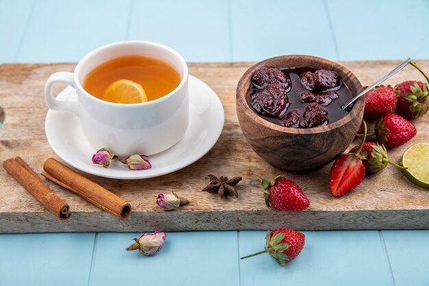 Vue de dessus de la confiture de fraises sur un bol en bois sur une planche de cuisine en bois avec une tasse de thé à la cannelle sur fond bleu