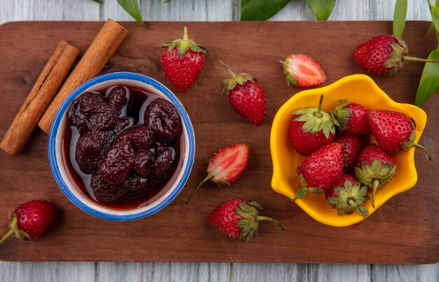 Vue de dessus de la confiture de fraises sur un bol sur une planche de cuisine en bois avec des fraises fraîches sur un bol jaune avec des bâtons de cannelle sur un fond en bois gris