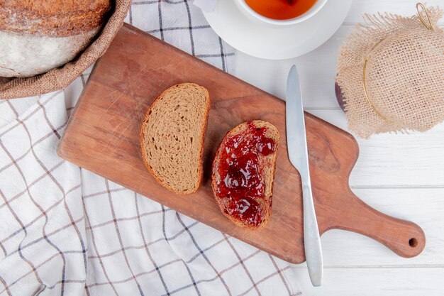 Vue de dessus de la confiture de fraises enduite de pain de seigle en tranches avec un couteau sur une planche à découper et des épis sur un tissu à carreaux avec du thé et de la confiture sur une table en bois