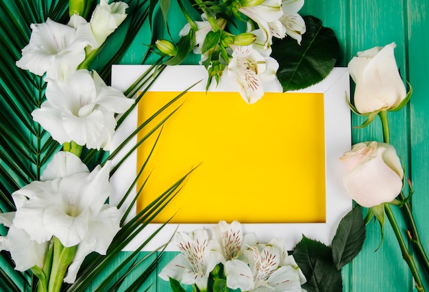 Photo gratuite vue de dessus de la couleur blanche alstroemeria et glaïeul avec feuille de palmier disposée autour d'un cadre avec une feuille de papier jaune sur fond vert