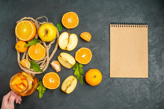 Vue de dessus coupé oranges et pommes bloc-notes cocktail dans une main féminine sur une surface sombre