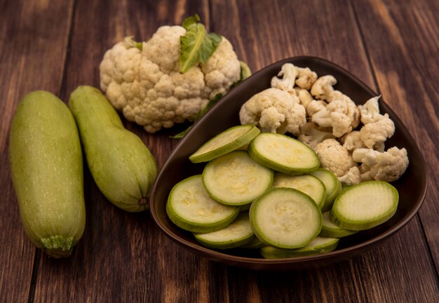 Vue de dessus des courgettes fraîches hachées et des bourgeons de chou-fleur sur un bol avec des courgettes entières et du chou-fleur isolé sur un mur en bois