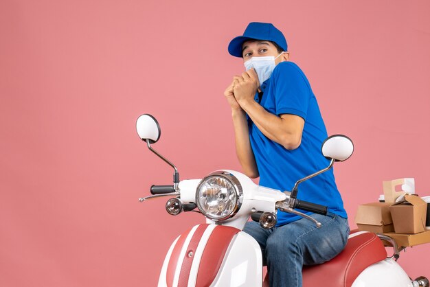 Vue de dessus d'un coursier effrayé portant un masque médical portant un chapeau assis sur un scooter livrant des commandes sur fond de pêche pastel