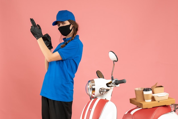 Vue de dessus d'une coursière confiante portant des gants de masque médical debout à côté d'une moto avec un gâteau au café dessus sur fond de couleur pêche pastel