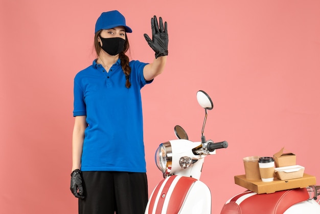 Vue de dessus d'une coursière confiante portant des gants de masque médical debout à côté d'une moto avec un gâteau au café dessus montrant cinq sur fond de couleur pêche pastel