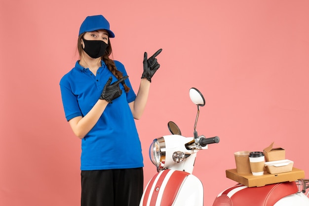 Vue de dessus d'une coursière confiante portant des gants de masque médical debout à côté d'une moto avec un gâteau au café pointant vers le haut sur un fond de couleur pêche pastel