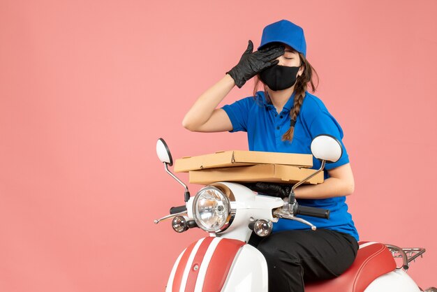 Vue de dessus d'une coursière fatiguée portant un masque médical et des gants assis sur un scooter livrant des commandes sur une pêche pastel