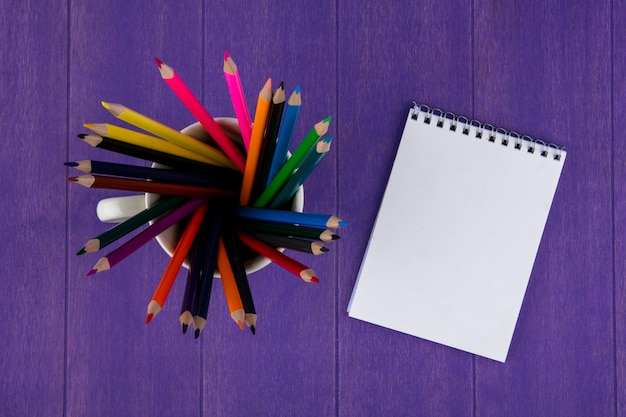 Vue de dessus des crayons de couleur en tasse et bloc-notes sur fond violet