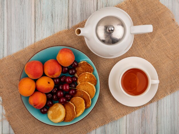 Vue de dessus des crêpes aux cerises et abricots en assiette et tasse de thé avec théière sur un sac et fond en bois