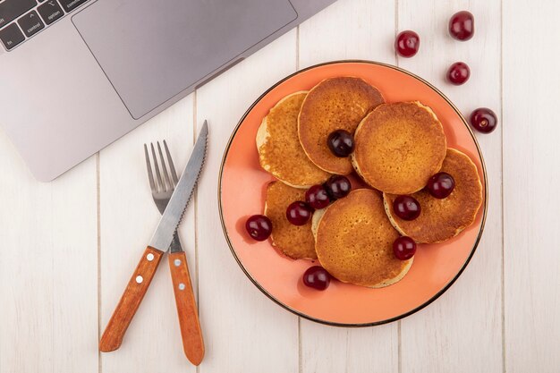 Vue de dessus des crêpes aux cerises en assiette et couteau fourchette et bloc-notes sur fond de bois