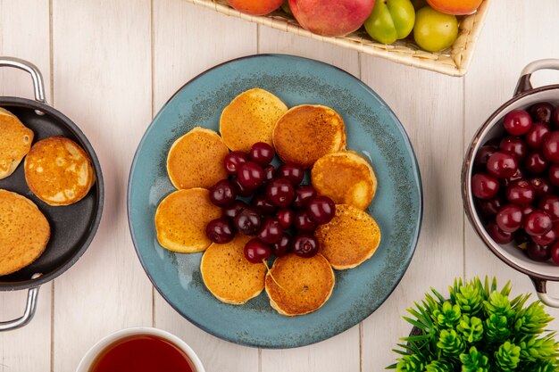 Vue de dessus des crêpes aux cerises en assiette et pan de crêpes avec bol de cerises et de fruits avec du thé sur fond de bois