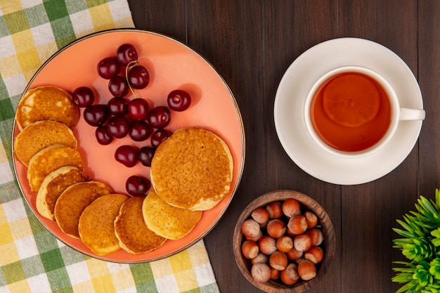 Vue de dessus des crêpes aux cerises en assiette sur tissu à carreaux et bol de noix avec tasse de thé sur fond de bois