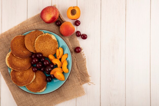 Vue de dessus des crêpes aux cerises et morceaux d'abricot en assiette et abricots cerises sur un sac et sur fond de bois avec espace copie