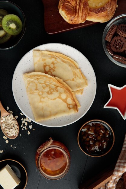 Vue de dessus des crêpes dans une assiette avec croissant kiwi biscuits confiture beurre d'avoine sur fond noir