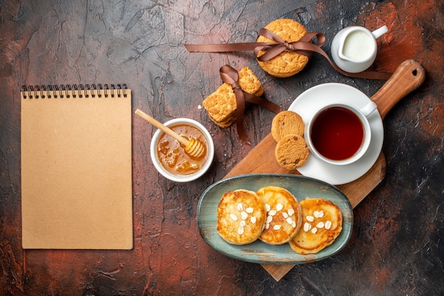 Vue de dessus de crêpes fraîches une tasse de thé noir sur une planche à découper en bois biscuits empilés au miel lait et cahier à spirale sur une surface sombre