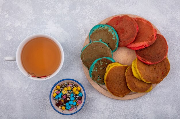 Vue de dessus des crêpes multicolores sur un support avec des chocolats colorés et une tasse de thé sur fond blanc