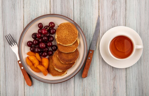 Vue de dessus des crêpes avec des tranches d'abricot et des cerises dans une assiette et une tasse de thé sur une soucoupe avec une fourchette et un couteau sur fond de bois