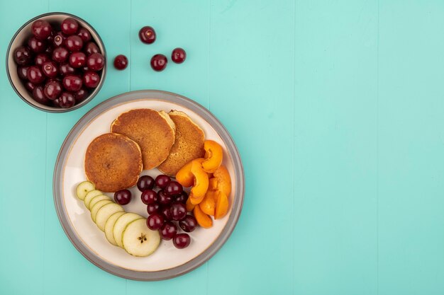 Vue de dessus des crêpes avec des tranches d'abricot et de poire et de cerises dans une assiette avec bol de cerises sur fond bleu avec copie espace