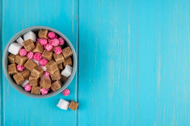 Vue de dessus des cubes de sucre brun et des bonbons roses dans un bol sur bleu