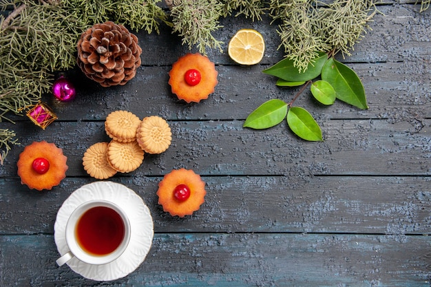 Vue de dessus cupcakes cerise branches de sapin tranche de citron une tasse de biscuits au thé et de feuilles sur une table en bois sombre avec espace copie