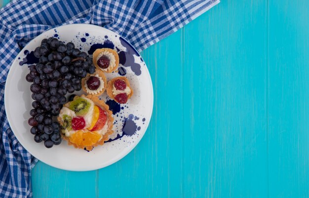 Vue de dessus des cupcakes fruités et du raisin en assiette sur un tissu à carreaux sur fond bleu avec espace copie