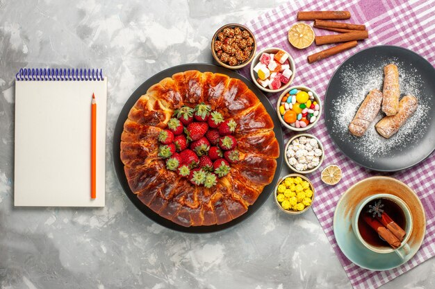 Vue de dessus de la délicieuse tarte aux fraises avec des bonbons aux fraises rouges fraîches et une tasse de thé sur une surface blanche