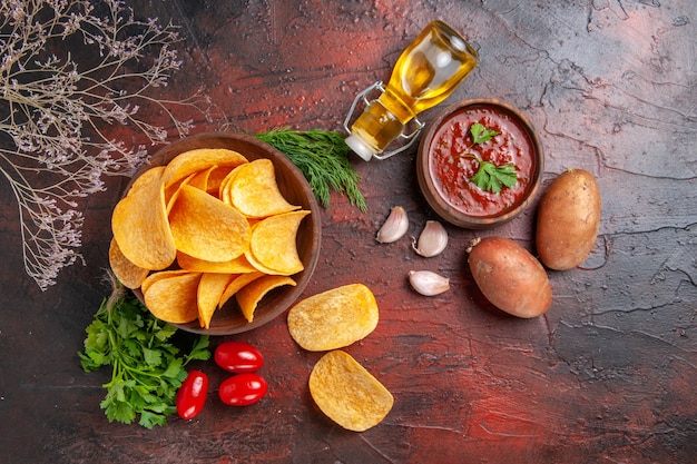 Vue de dessus de délicieuses chips croustillantes de pommes de terre faites maison dans un petit bol brun bouteille d'huile de pommes de terre tomates vertes ail et ketchup sur fond sombre