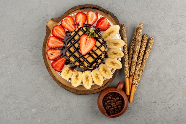 Vue de dessus de délicieuses crêpes sucrées délicieuses avec des fraises rouges en tranches et des bananes à l'intérieur de la plaque sur le fond gris