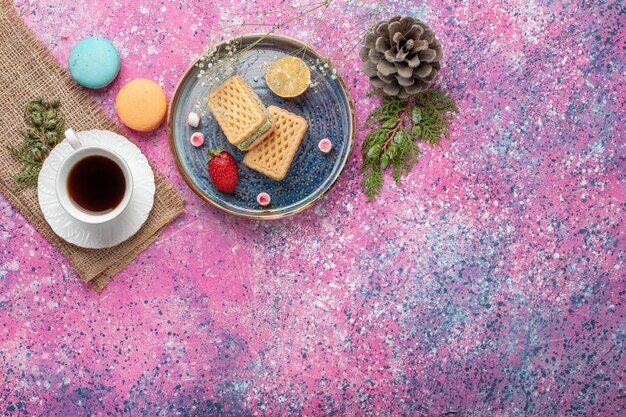 Vue de dessus de délicieuses gaufres avec tasse de thé et macarons sur surface rose