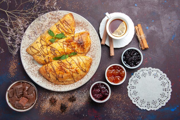 Vue de dessus de délicieuses pâtisseries cuites au four avec tasse de confiture de thé sur un espace sombre