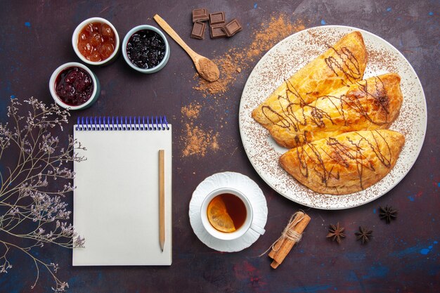 Vue de dessus de délicieuses pâtisseries cuites au four avec tasse de confiture de thé sur un espace sombre