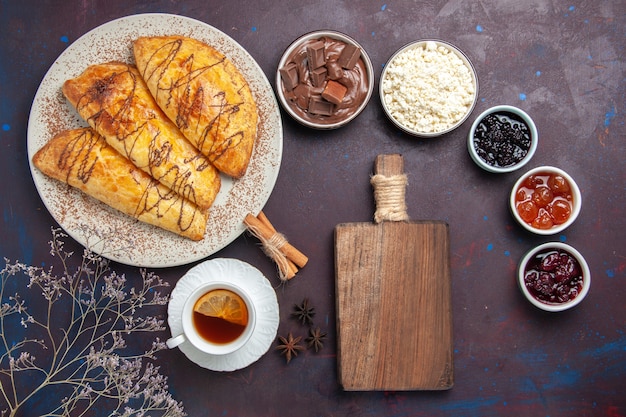 Vue de dessus de délicieuses pâtisseries cuites au four avec une tasse de thé et de confiture sur un bureau sombre