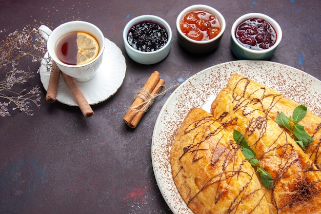 Vue de dessus de délicieuses pâtisseries cuites au four avec une tasse de thé et de confiture sur un bureau sombre