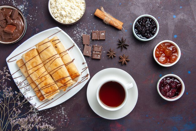 Vue de dessus de délicieuses pâtisseries sucrées avec de la confiture de chocolat et une tasse de thé sur un espace sombre