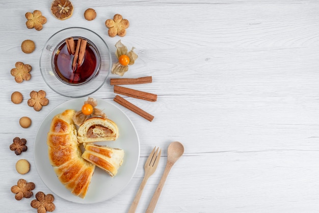 Vue de dessus de délicieuses pâtisseries en tranches à l'intérieur de la plaque avec remplissage avec du thé et des biscuits sur un bureau blanc, biscuit de pâtisserie biscuit sucré