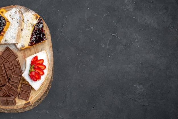 Vue de dessus de délicieuses tranches de gâteau avec des fraises et des barres de chocolat sur une surface sombre