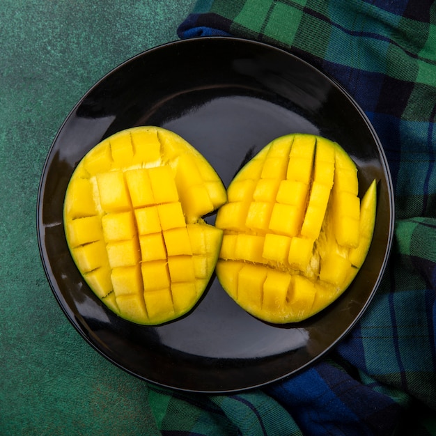 Vue de dessus de délicieuses tranches de mangue fraîche jaune dans une plaque noire sur nappe à carreaux et surface verte