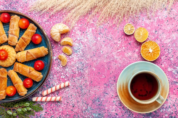 Vue de dessus de délicieux bagels sucrés à l'intérieur du plateau avec des prunes aigres et du thé sur le bureau rose clair