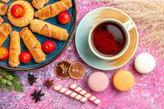 Vue de dessus de délicieux bagels sucrés à l'intérieur du plateau avec des prunes aigres fraîches macarons français et tasse de thé sur un bureau rose clair