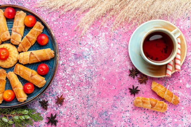 Vue de dessus de délicieux bagels sucrés à l'intérieur du plateau avec des prunes aigres fraîches et une tasse de thé sur un bureau rose clair