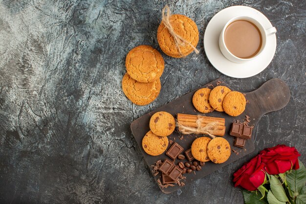 Vue de dessus de délicieux biscuits barres de chocolat roses rouges et une tasse de café sur le côté gauche sur fond sombre glacial